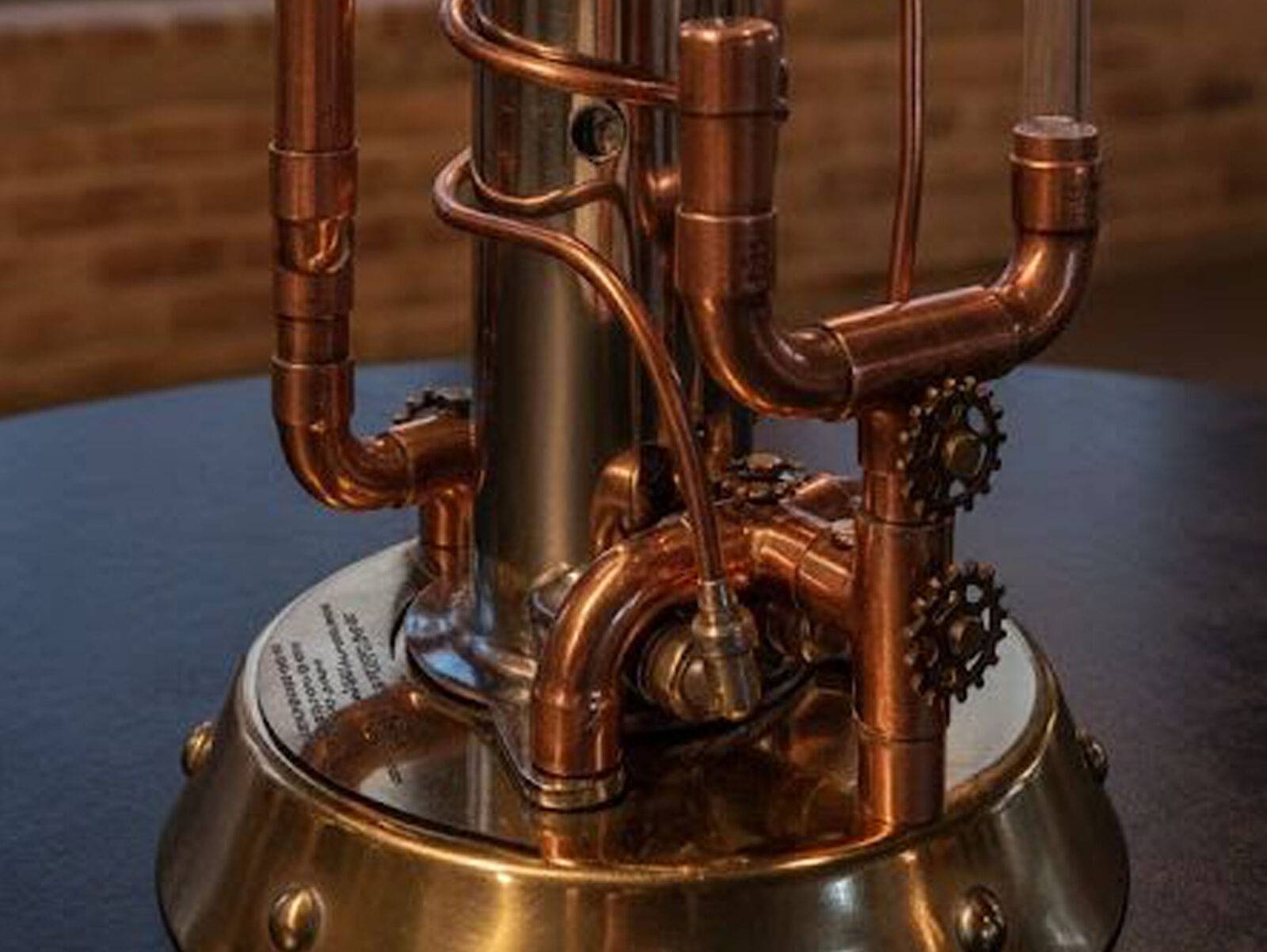 Pionowa rzeźba steampunkowa wykonana z miedzianych rur, mosiężnych zębatek, nitowanej podstawy i centralnego cylindra ze stali nierdzewnej, przypominająca retro-futurystyczny destylator